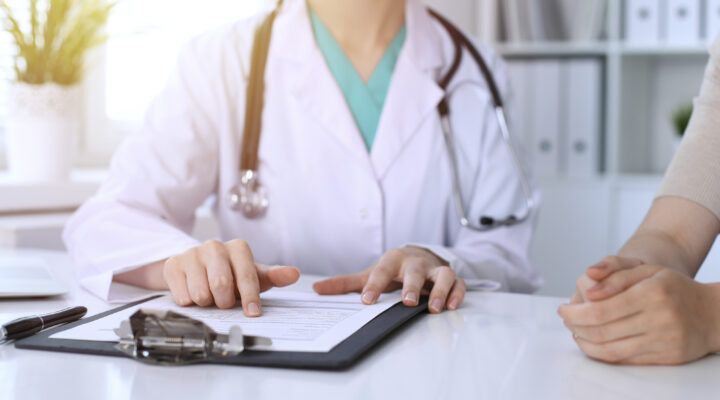 Ärztin oder Arzt im weißen Kittel erklärt einer Person ein Dokument auf einem Klemmbrett und zeigt mit dem Finger auf einen Abschnitt; gegenüber sitzen die gefalteten Hände der zu beratenden Person auf dem Tisch.