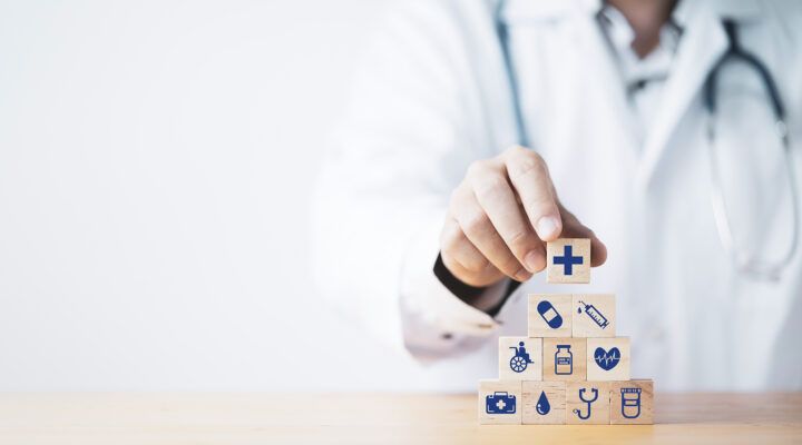 Person im weißen Arztkittel mit Stethoskop stapelt Holzklötze mit medizinischen Symbolen wie Kreuz, Spritze, Herz, Rollstuhl und Medikamenten.