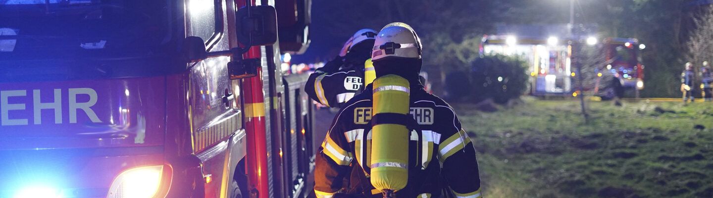 Feuerwehrleute in voller Schutzausrüstung mit Atemgeräten gehen nachts an einem Einsatzfahrzeug vorbei, im Hintergrund weitere beleuchtete Feuerwehrfahrzeuge und Einsatzkräfte auf einer Wiese.