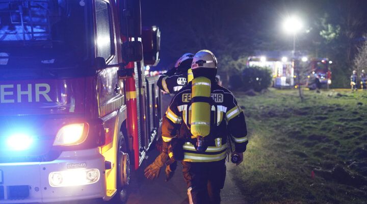 Feuerwehrleute in voller Schutzausrüstung mit Atemgeräten gehen nachts an einem Einsatzfahrzeug vorbei, im Hintergrund weitere beleuchtete Feuerwehrfahrzeuge und Einsatzkräfte auf einer Wiese.