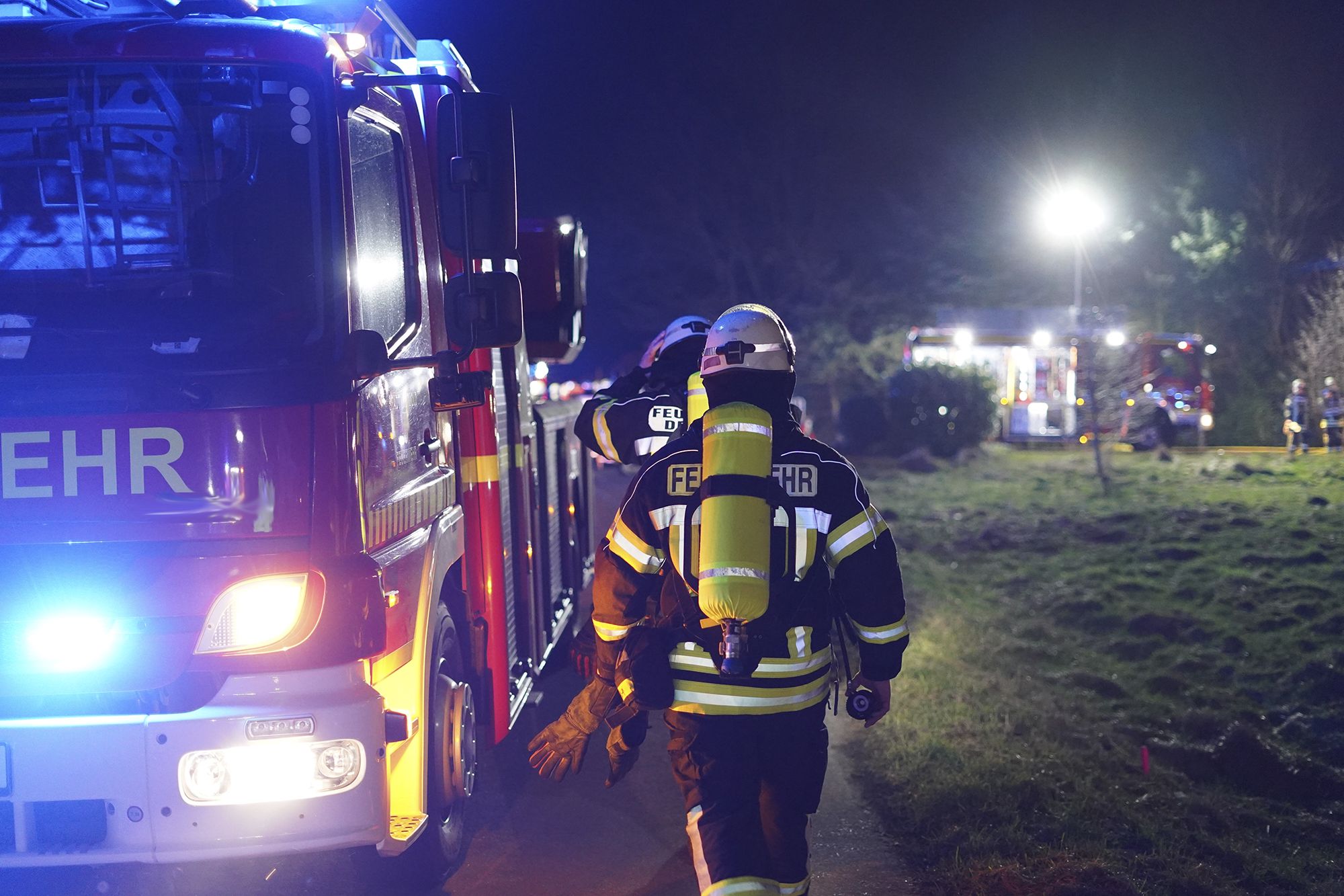 Feuerwehrleute in voller Schutzausrüstung mit Atemgeräten gehen nachts an einem Einsatzfahrzeug vorbei, im Hintergrund weitere beleuchtete Feuerwehrfahrzeuge und Einsatzkräfte auf einer Wiese.