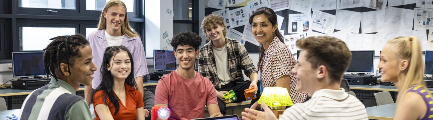 Eine Gruppe Jugendlicher sitzt gemeinsam in einem Klassenraum vor Computern und unterhält sich lachend. Einige halten kleine technische Modelle oder Bauteile in den Händen, während sie im Kreis zusammensitzen und diskutieren.