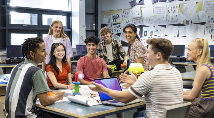 Eine Gruppe Jugendlicher sitzt gemeinsam in einem Klassenraum vor Computern und unterhält sich lachend. Einige halten kleine technische Modelle oder Bauteile in den Händen, während sie im Kreis zusammensitzen und diskutieren.