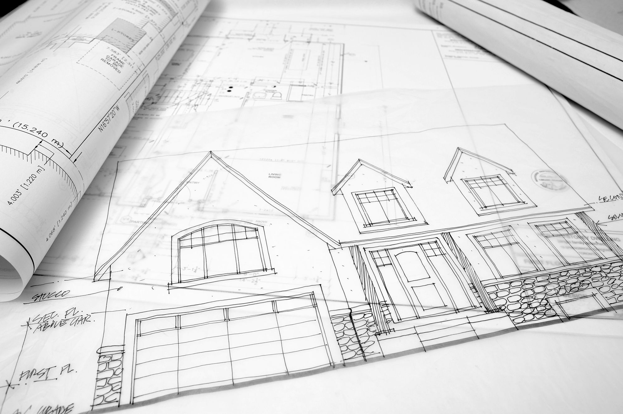 Schwarz-weißes Foto von ausgebreiteten Bauplänen auf einem Tisch. Im Vordergrund ist eine detaillierte Architekturzeichnung der Fassade eines Einfamilienhauses mit Garage, Haustür und Dachgauben zu sehen. Im Hintergrund liegen weitere aufgerollte Pläne und technische Zeichnungen mit Maßangaben.