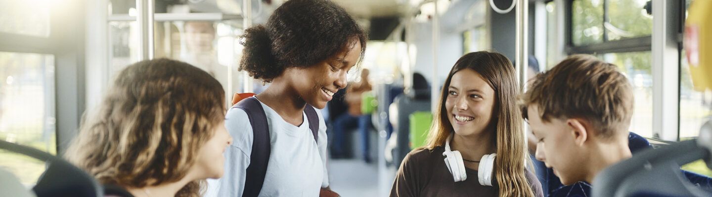 Vier Jugendliche stehen gemeinsam in einem Bus und unterhalten sich fröhlich. Zwei Mädchen und zwei Jungen lächeln sich an, eine trägt Kopfhörer um den Hals, ein anderes hat einen Rucksack auf. Im Hintergrund sind Bussitze und Haltestangen zu sehen, Tageslicht fällt durch die Fenster.