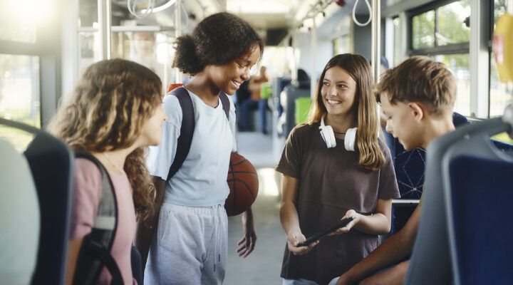 Vier Jugendliche stehen gemeinsam in einem Bus und unterhalten sich fröhlich. Zwei Mädchen und zwei Jungen lächeln sich an, eine trägt Kopfhörer um den Hals, ein anderes hat einen Rucksack auf. Im Hintergrund sind Bussitze und Haltestangen zu sehen, Tageslicht fällt durch die Fenster.