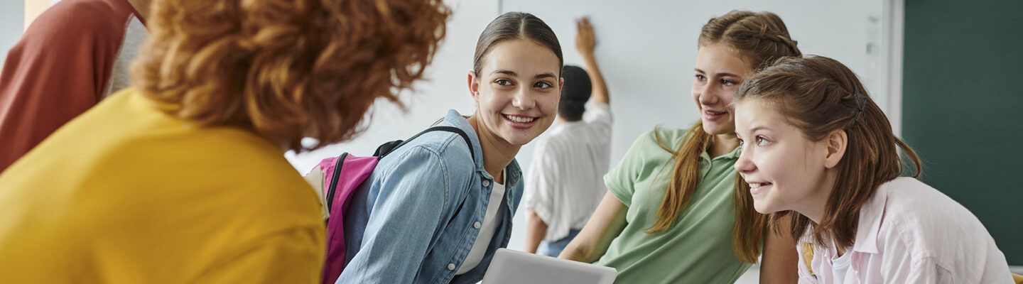 Drei Schülerinnen stehen in einem Klassenraum zusammen und unterhalten sich lächelnd mit einer erwachsenen Person im Vordergrund. Eine der Schülerinnen trägt einen Rucksack und hält ein Tablet in der Hand. Im Hintergrund ist eine Tafel zu sehen, an der eine weitere Person schreibt.
