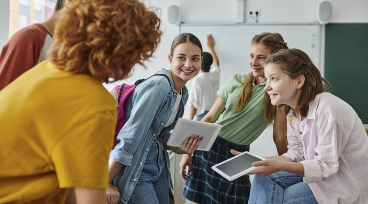 Drei Schülerinnen stehen in einem Klassenraum zusammen und unterhalten sich lächelnd mit einer erwachsenen Person im Vordergrund. Eine der Schülerinnen trägt einen Rucksack und hält ein Tablet in der Hand. Im Hintergrund ist eine Tafel zu sehen, an der eine weitere Person schreibt.