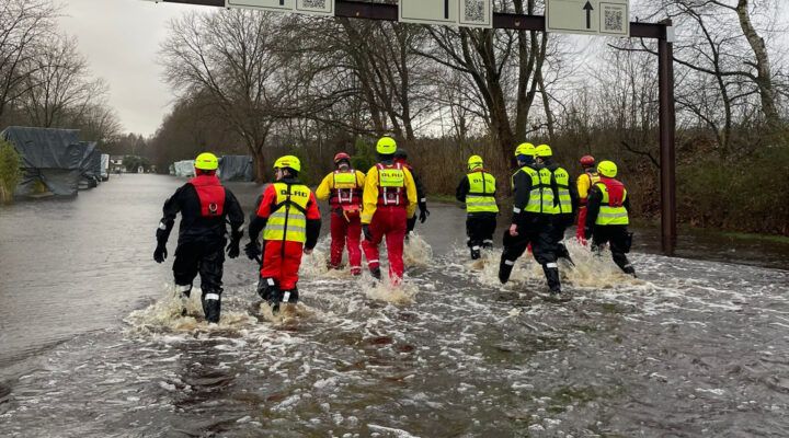 Einsatzkräfte in Schutzanzügen und Warnwesten waten durch eine überflutete Straße. Im Hintergrund sind Bäume und Einsatzfahrzeuge zu sehen, die auf einen Hochwassereinsatz hinweisen.