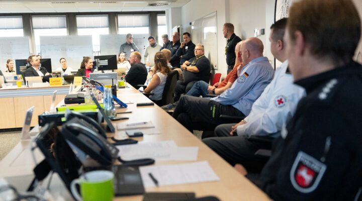 Katastrophenschutzübung: Mehrere Einsatzkräfte und Mitarbeitende verschiedener Organisationen sitzen und stehen in einem Besprechungsraum. Auf den Tischen liegen Unterlagen, Telefone und Computer. Im Hintergrund sind Whiteboards mit Notizen zu sehen.