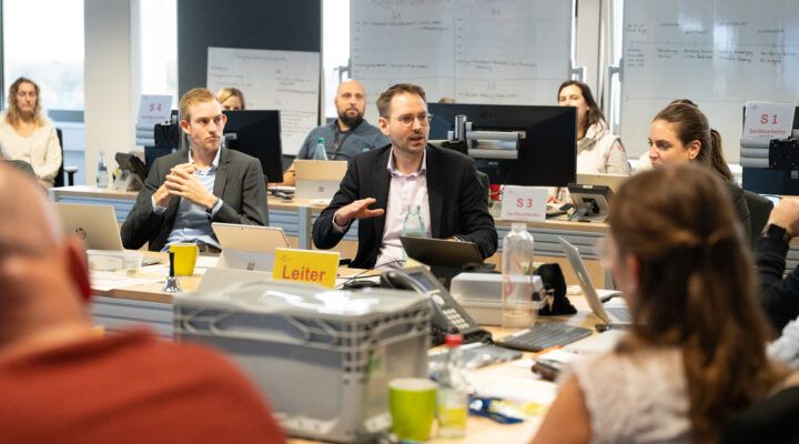 Mehrere Personen sitzen in einem Besprechungsraum mit Computern und Whiteboards. In der Mitte spricht Stabsleiter Sebastian Smolinski,. Die Gruppe wirkt konzentriert und befindet sich in einer Lagebesprechung.