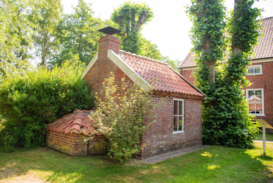 Kleines Backsteingebäude mit rotem Ziegeldach und Schornstein steht in einem sonnigen Garten, umgeben von Büschen, Bäumen und gepflegtem Rasen.