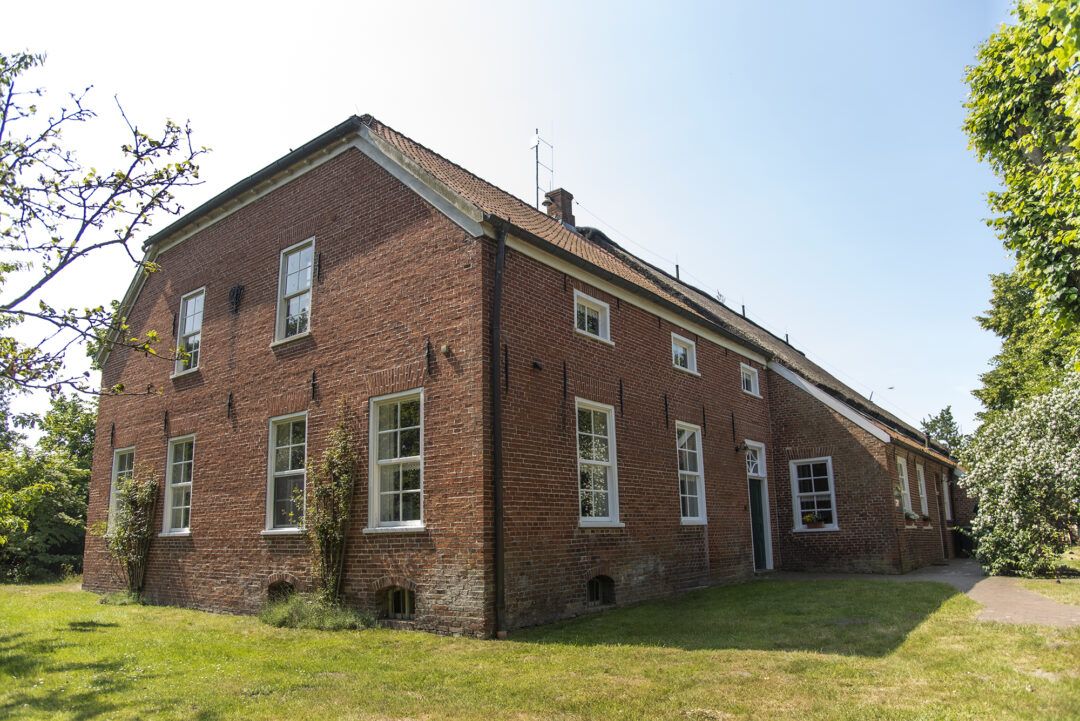 Zweigeschossiges, historisches Backsteingebäude mit Satteldach und weißen Sprossenfenstern, umgeben von Rasen und Bäumen bei sonnigem Wetter.
