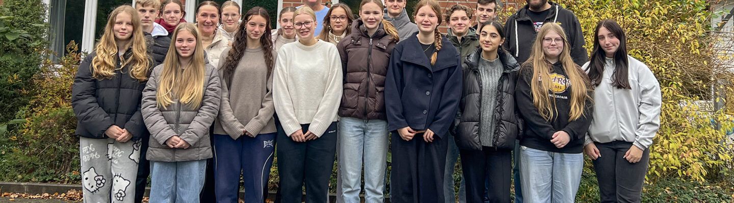 Eine Gruppe von Jugendlichen und zwei erwachsenen Begleitpersonen steht in mehreren Reihen vor einem Backsteingebäude. Alle blicken in die Kamera und tragen herbstliche Kleidung. Über dem Eingang ist ein Schild mit dem DJH-Logo zu sehen, im Hintergrund befinden sich Büsche und Bäume.