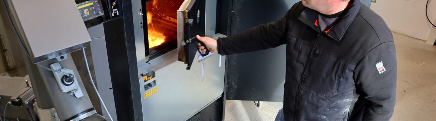 Eine Person bedient eine industrielle Heizungs- oder Verbrennungsanlage in einem Technikraum und öffnet eine Feuerkammer, in der Flammen sichtbar sind. Daneben befinden sich Rohre, Steuerungselemente und technische Geräte.