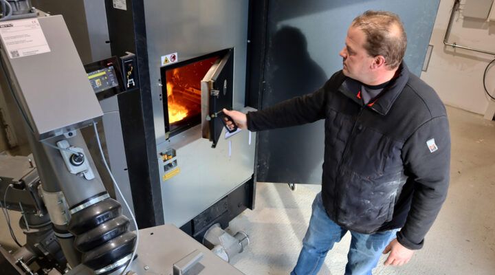 Eine Person bedient eine industrielle Heizungs- oder Verbrennungsanlage in einem Technikraum und öffnet eine Feuerkammer, in der Flammen sichtbar sind. Daneben befinden sich Rohre, Steuerungselemente und technische Geräte.