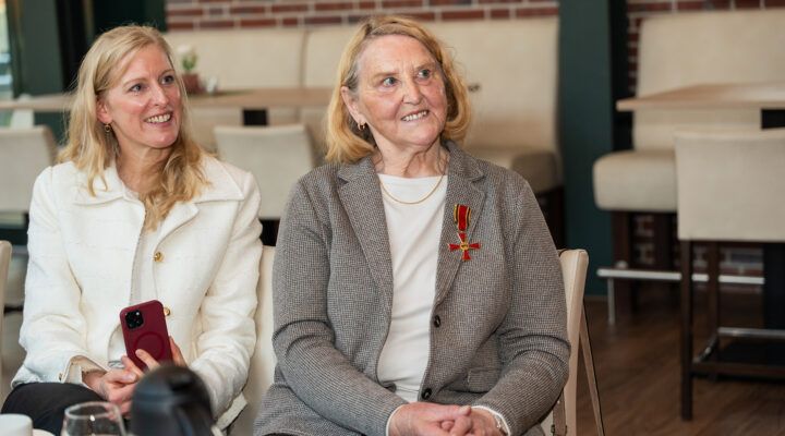 Anna Uphoff und ihre Tochter sitzen in einem modern eingerichteten Raum nebeneinander und lächeln. Anna Uphoff trägt ein Jackett und das Bundesverdienstkreuzes an ihrer Brust; die Tochter links hält ein Smartphone in der Hand. Im Hintergrund stehen helle Stühle und Tische.