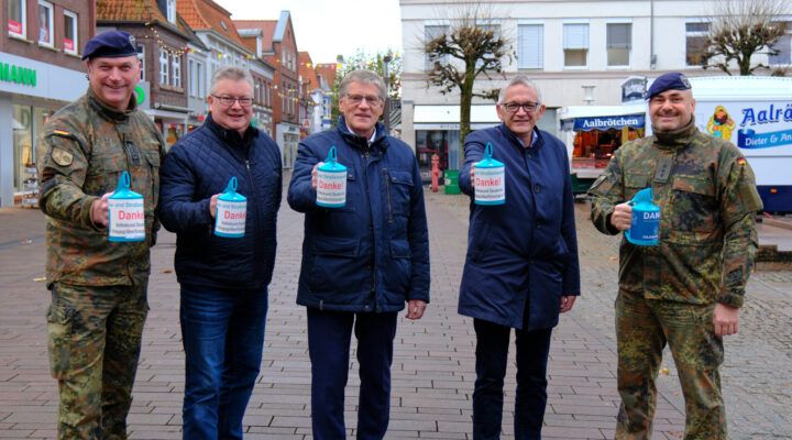 Fünf Männer stehen in einer Einkaufsstraße und halten jeweils eine Sammeldose mit der Aufschrift ‚Danke!‘ in der Hand. Zwei der Männer tragen Bundeswehruniformen, die drei anderen dunkle Jacken. Im Hintergrund sind Geschäfte und ein Brötchenwagen zu sehen.