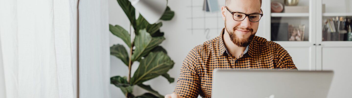 Ein Mann sitzt an einem hellen Schreibtisch in einem modernen Arbeitszimmer und arbeitet an einem Laptop. Neben ihm stehen eine Tischlampe, Notizunterlagen und ein Smartphone; im Hintergrund sind Pflanzen und ein Regal mit Büchern zu sehen.