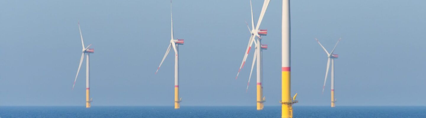 Mehrere Offshore-Windkraftanlagen stehen im Meer; ihre weißen Rotorblätter drehen sich vor einem klaren blauen Himmel, die Fundamente ragen gelb aus dem Wasser.