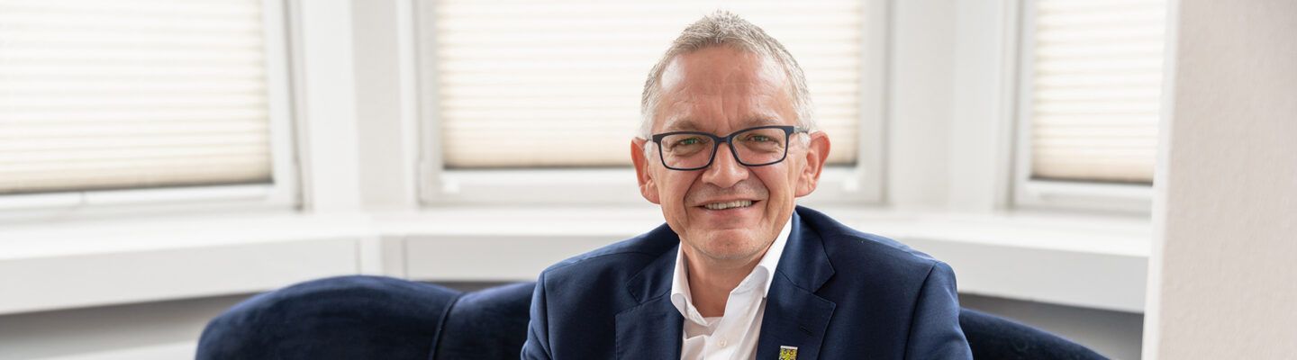 Landrat Olaf Meinen sitzt lächelnd auf einem roten Sofa in einem hellen Raum. Er trägt einen dunkelblauen Anzug mit weißem Hemd und Brille; hinter ihm sind Fenster mit Jalousien zu sehen.