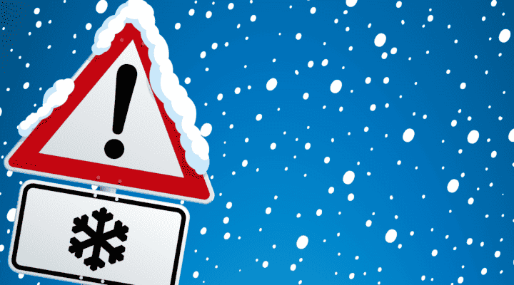 Verkehrszeichen mit rotem Warn-Dreieck und Ausrufezeichen, darunter ein Zusatzschild mit Schneeflocken-Symbol. Das Schild ist mit Schnee bedeckt, im Hintergrund fällt dichter Schnee vor blauem Himmel – Hinweis auf winterliche Glätte.