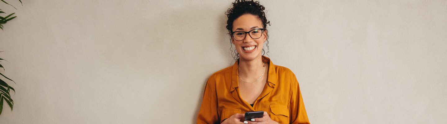 „Eine Frau steht lächelnd vor einer hellen Wand und hält ein Smartphone mit beiden Händen. Sie trägt eine orangefarbene Bluse, eine Jeans und eine Brille; links ist eine Zimmerpflanze angeschnitten zu sehen.