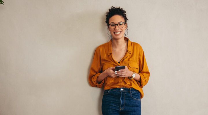 „Eine Frau steht lächelnd vor einer hellen Wand und hält ein Smartphone mit beiden Händen. Sie trägt eine orangefarbene Bluse, eine Jeans und eine Brille; links ist eine Zimmerpflanze angeschnitten zu sehen.