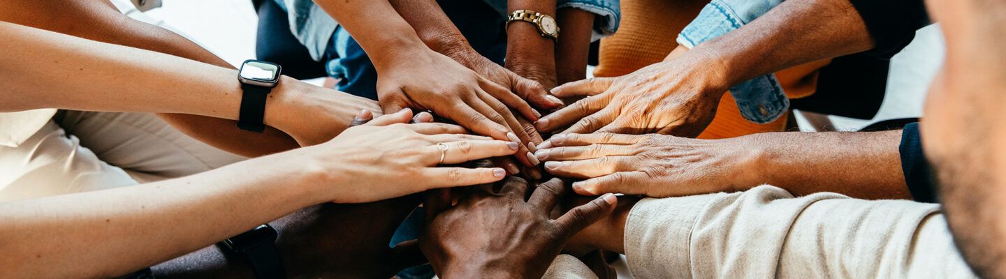 Mehrere Menschen unterschiedlicher Hautfarben legen ihre Hände übereinander in der Mitte eines Kreises. Die Aufnahme symbolisiert Gemeinschaft, Zusammenhalt und Teamarbeit.