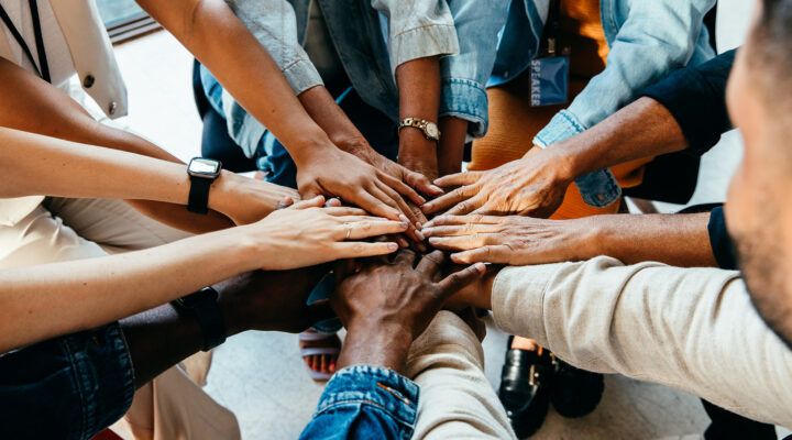 Mehrere Menschen unterschiedlicher Hautfarben legen ihre Hände übereinander in der Mitte eines Kreises. Die Aufnahme symbolisiert Gemeinschaft, Zusammenhalt und Teamarbeit.