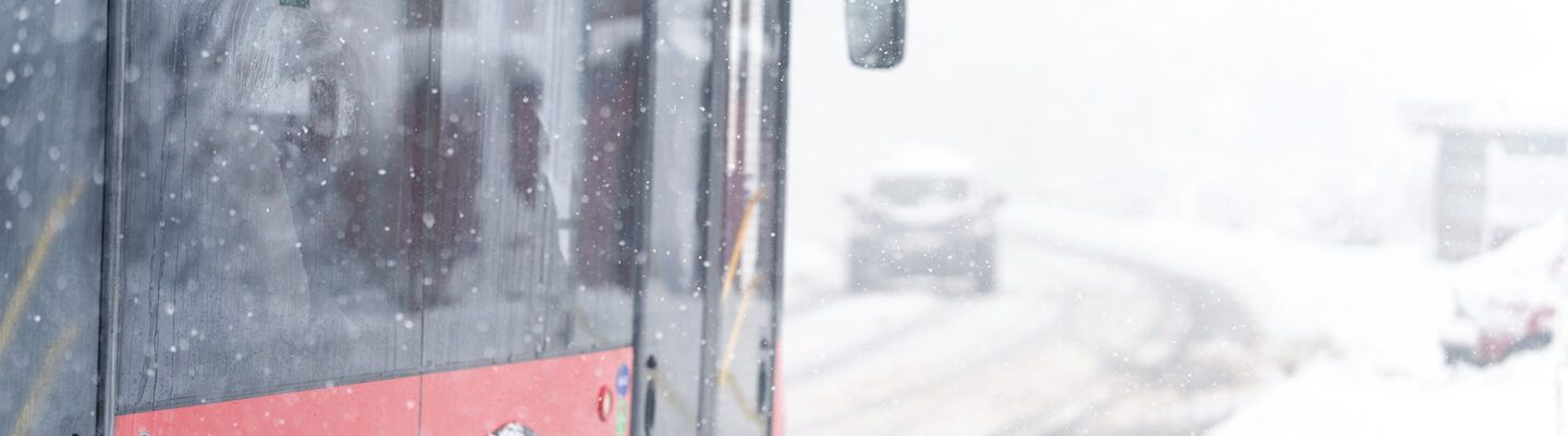 Ein Linienbus fährt bei starkem Schneefall auf einer verschneiten Straße. Schneeflocken und aufgewirbelter Schnee schränken die Sicht ein, die Fahrbahn ist winterlich bedeckt.