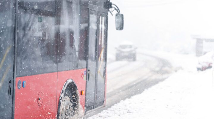 Ein Linienbus fährt bei starkem Schneefall auf einer verschneiten Straße. Schneeflocken und aufgewirbelter Schnee schränken die Sicht ein, die Fahrbahn ist winterlich bedeckt.