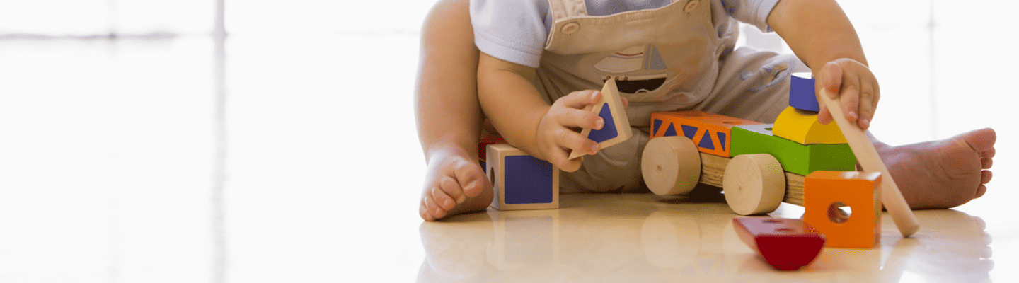 Kleinkind sitzt auf dem Boden und spielt mit bunten Holzbausteinen und einem Holzfahrzeug. Das Kind hält einen Baustein in der Hand, im Hintergrund fällt helles Tageslicht durch große Fenster.