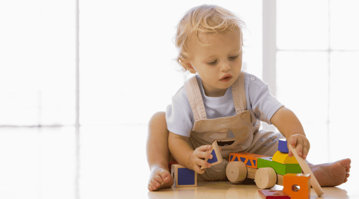 Kleinkind sitzt auf dem Boden und spielt mit bunten Holzbausteinen und einem Holzfahrzeug. Das Kind hält einen Baustein in der Hand, im Hintergrund fällt helles Tageslicht durch große Fenster.