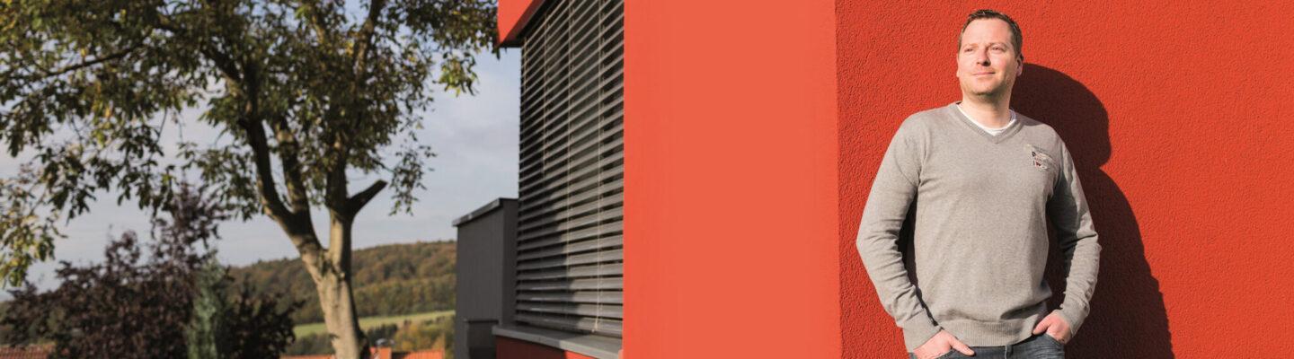 Eine Person steht an der Außenwand eines modernen, rot gestrichenen Gebäudes und blickt seitlich in die Sonne. An der Wand ist ein grünes Schild mit der Zahl 27 angebracht. Im Hintergrund sind Bäume und eine ländliche Landschaft zu sehen.