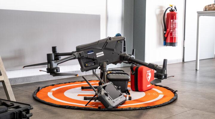 Große Drohne steht auf einer orangefarbenen Landeplattform in einem Innenraum; davor liegt eine Fernsteuerung. Im Hintergrund sind ein roter Feuerlöscher und eine Tasche mit Erste-Hilfe-Symbol zu sehen.