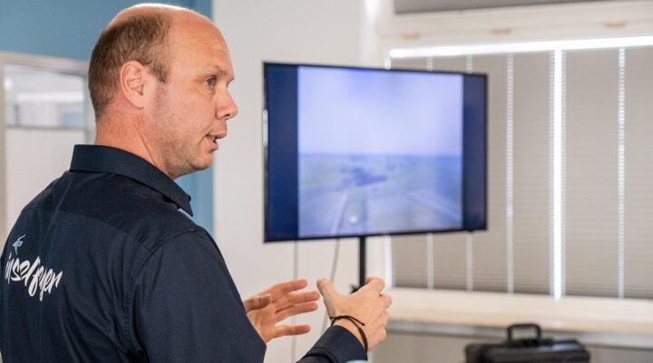 Ein Mann erklärt etwas mit Gesten vor einem Bildschirm, auf dem eine Luftaufnahme zu sehen ist, vermutlich von einer Drohne.