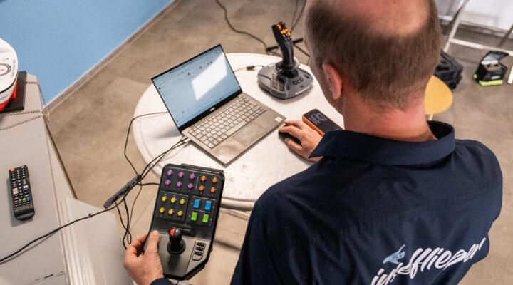 Eine Person sitzt an einem Tisch und steuert mit einem Bedienpult eine Drohne; vor ihr stehen ein Laptop und eine weitere Fernsteuerung.