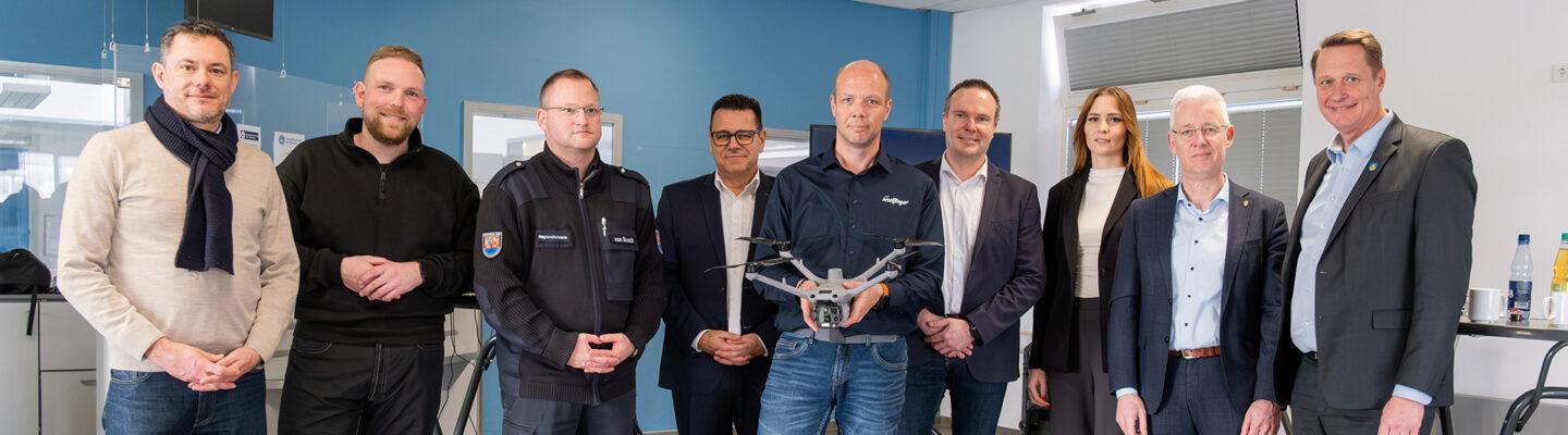 Eine Gruppe von neun Erwachsenen steht in einem Innenraum nebeneinander; eine Person in der Mitte hält eine Drohne. Vor ihnen steht ein dreieckiges Warnschild mit der Aufschrift „Achtung Drohnenflug“.