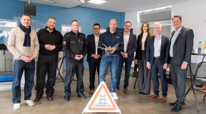 Eine Gruppe von neun Erwachsenen steht in einem Innenraum nebeneinander; eine Person in der Mitte hält eine Drohne. Vor ihnen steht ein dreieckiges Warnschild mit der Aufschrift „Achtung Drohnenflug“.