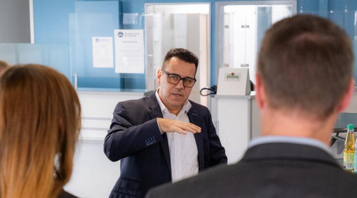 Ein Mann im Anzug erklärt etwas gestikulierend in einem Raum; zwei Personen hören ihm im Vordergrund zu.