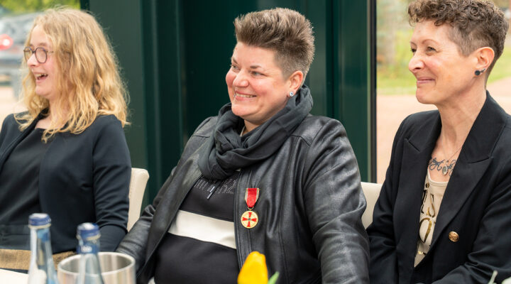 Tochter Juna, Melanie Doden und Ehefrau Ingrid Doden sitzen nebeneinander an einem Tisch und lächeln; Melane Doden in der Mitte trägt eine Auszeichnung an einer roten Schleife.