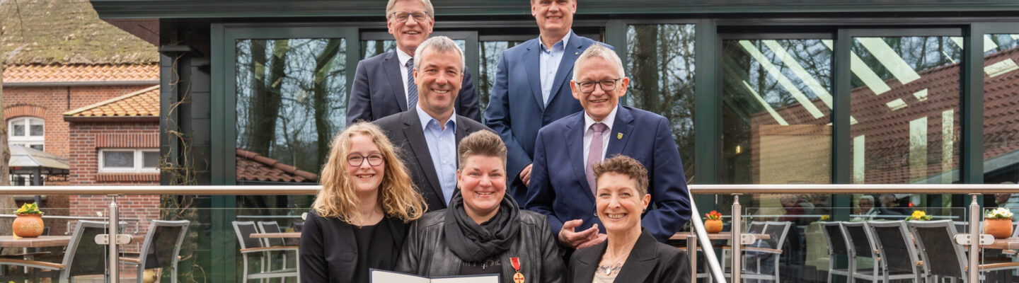 Gruppe von sieben Personen steht vor einem Gebäude; in der Mitte hält Frau Melanie Doden eine Urkunde und einen Blumenstrauß.