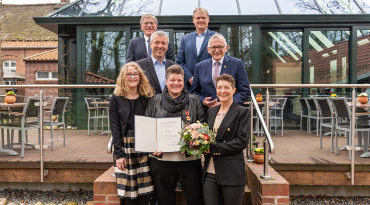 Gruppe von sieben Personen steht vor einem Gebäude; in der Mitte hält Frau Melanie Doden eine Urkunde und einen Blumenstrauß.