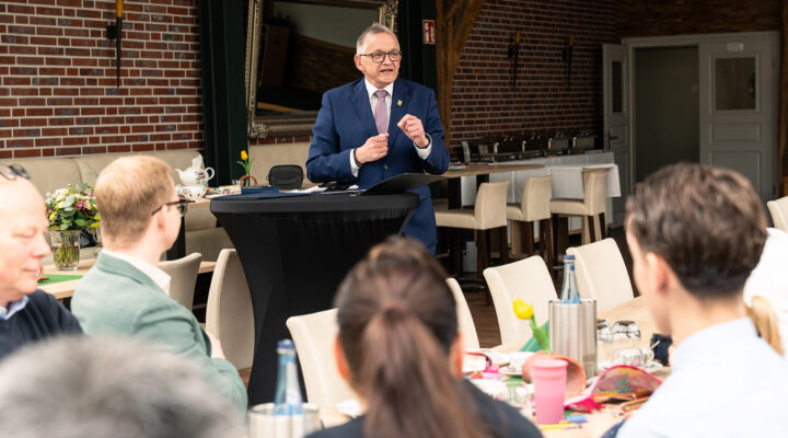Landrat Olaf Meinen steht an einem Stehtisch und hält eine Rede vor mehreren Zuhörenden in einem Veranstaltungsraum mit Backsteinwänden und gedeckten Tischen.