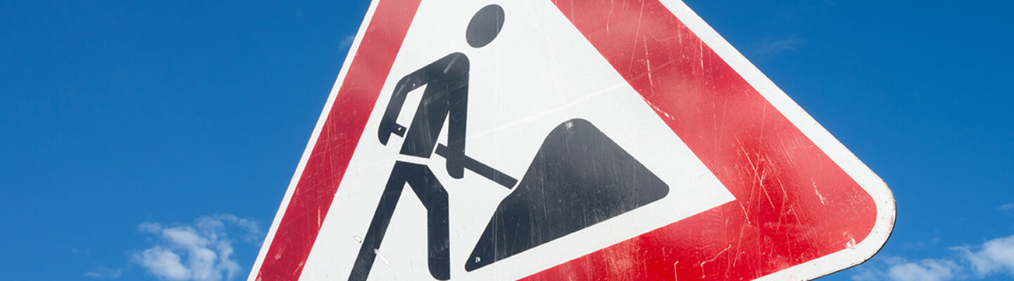 Dreieckiges Verkehrsschild mit rotem Rand und Baustellensymbol (arbeitende Person mit Schaufel) vor blauem Himmel.