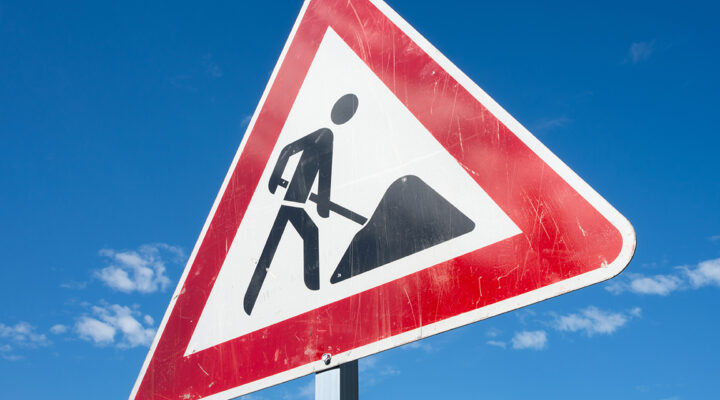 Dreieckiges Verkehrsschild mit rotem Rand und Baustellensymbol (arbeitende Person mit Schaufel) vor blauem Himmel.
