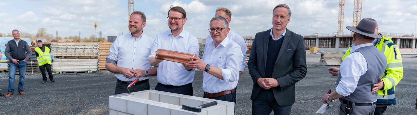 Tim Kruithoff, Oberbürgermeister der Stadt Emden und Aufsichtsratsvorsitzender der Trägergesellschaft Kliniken Aurich-Emden-Norden mbH, Dirk Balster, Geschäftsführer der Trägergesellschaft Kliniken Aurich-Emden-Norden mbH, Olaf Meinen, Landrat des Landkreises Aurich, Matthias Arends, Landtagsabgeordneter (MdL) und Wiard Siebels, Landtagsabgeordneter (MdL) stehen auf einer Baustelle um ein gemauertes Fundament; zwei von ihnen halten eine Kupferkapsel für eine symbolische Grundsteinlegung, während andere zuschauen.