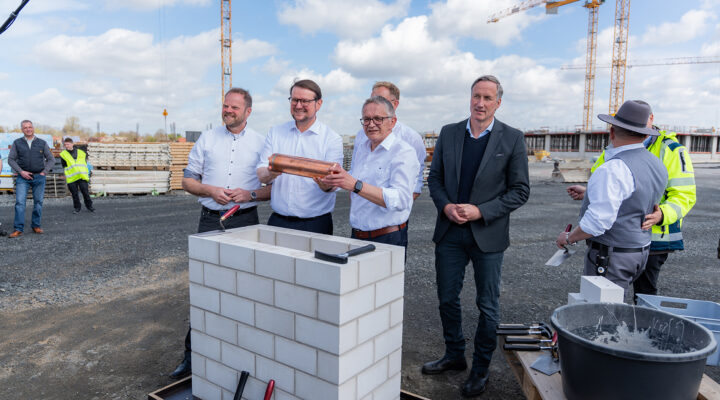 Tim Kruithoff, Oberbürgermeister der Stadt Emden und Aufsichtsratsvorsitzender der Trägergesellschaft Kliniken Aurich-Emden-Norden mbH, Dirk Balster, Geschäftsführer der Trägergesellschaft Kliniken Aurich-Emden-Norden mbH, Olaf Meinen, Landrat des Landkreises Aurich, Matthias Arends, Landtagsabgeordneter (MdL) und Wiard Siebels, Landtagsabgeordneter (MdL) stehen auf einer Baustelle um ein gemauertes Fundament; zwei von ihnen halten eine Kupferkapsel für eine symbolische Grundsteinlegung, während andere zuschauen.