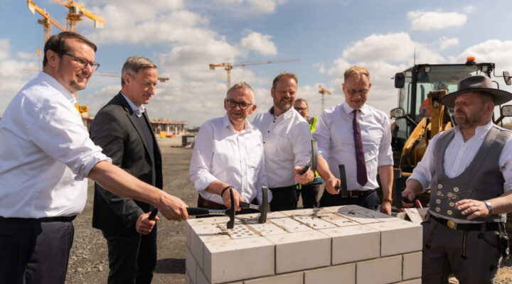 Tim Kruithoff, Oberbürgermeister der Stadt Emden und Aufsichtsratsvorsitzender der Trägergesellschaft Kliniken Aurich-Emden-Norden mbH, Dirk Balster, Geschäftsführer der Trägergesellschaft Kliniken Aurich-Emden-Norden mbH, Olaf Meinen, Landrat des Landkreises Aurich, Matthias Arends, Landtagsabgeordneter (MdL) und Wiard Siebels, Landtagsabgeordneter (MdL) stehen um ein gemauertes Fundament und schlagen gemeinsam mit Hämmern symbolisch auf die Steine; im Hintergrund sind Kräne und Baumaschinen zu sehen.